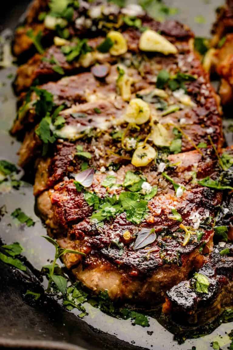 Close-up of a cooked steak topped with sliced garlic, chopped fresh herbs, and coarse salt, served on a dark surface. The steak appears juicy and well-seasoned.