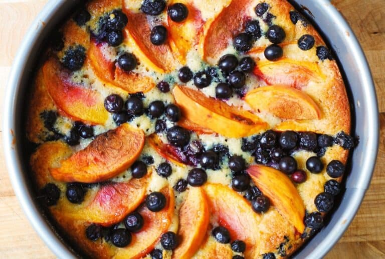 A round cake topped with slices of peach and scattered blueberries, baked to a golden brown in a metal pan, sits on a wooden surface.
