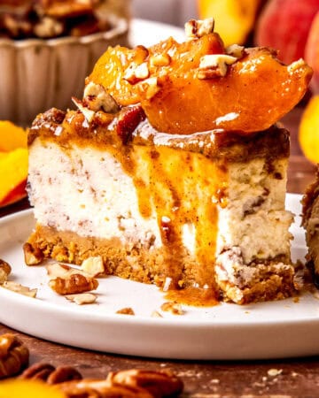 A slice of creamy peach cobbler cheesecake topped with peach slices, chopped pecans, and sauce sits on a white plate.
