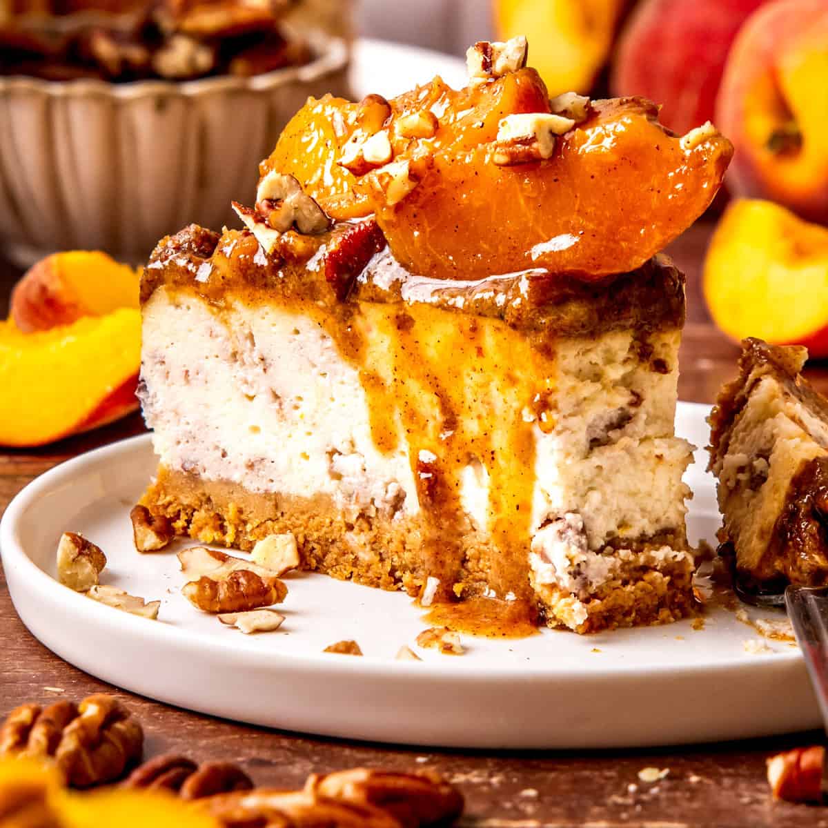 A slice of creamy peach cobbler cheesecake topped with peach slices, chopped pecans, and sauce sits on a white plate. 