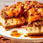 A close-up of a peach cobbler cheesecake topped with caramelized peaches and chopped nuts, set on a white cake stand. A slice has been removed, revealing the creamy filling and crumbly crust.