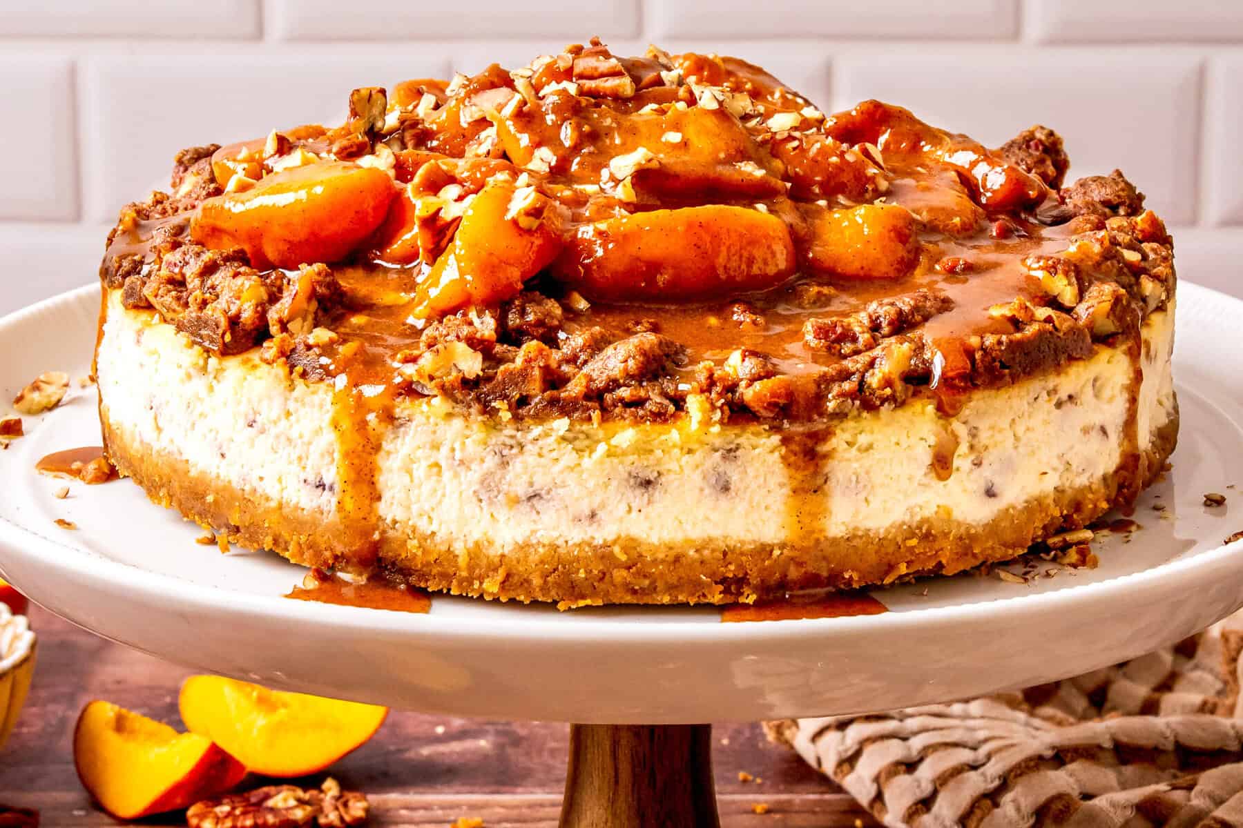 A peach cobbler cheesecake topped with caramelized peaches and chopped pecans sits on a white cake stand, with sauce dripping down the sides. The dessert is set against a tiled white background.
