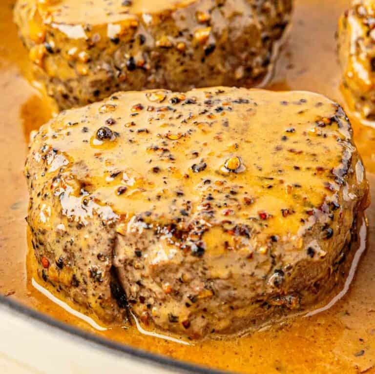 A close-up of a filet mignon steak covered in a creamy peppercorn sauce, showing a juicy texture and visible cracked pepper and spice. The steak is sitting in a pool of sauce in a pan.
