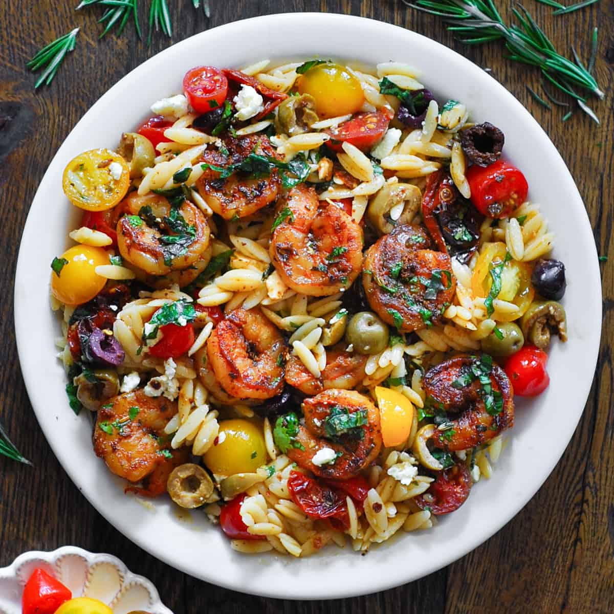 A white plate filled with orzo pasta, seared shrimp, cherry tomatoes, olives, and herbs, garnished with crumbled feta cheese, sits on a wooden table with sprigs of rosemary nearby.