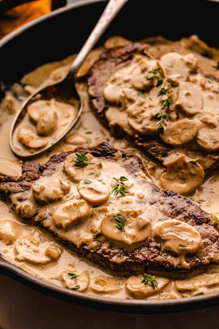 Two cooked steaks in a skillet, topped with creamy mushroom sauce and garnished with fresh herbs, with a spoon resting in the sauce.