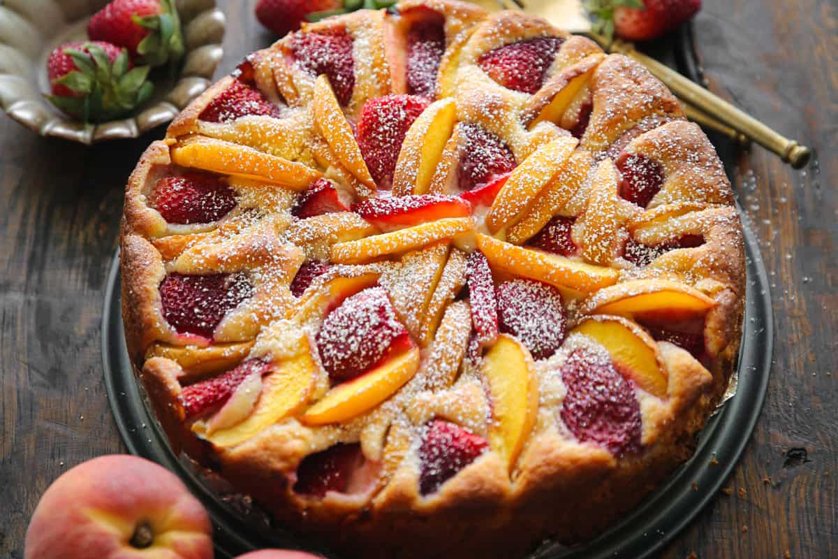 These 10 Peach Desserts Are Almost Too Pretty to Eat A round strawberry peach cake topped with slices of fresh peaches and strawberries, dusted with powdered sugar, sits on a dark plate.