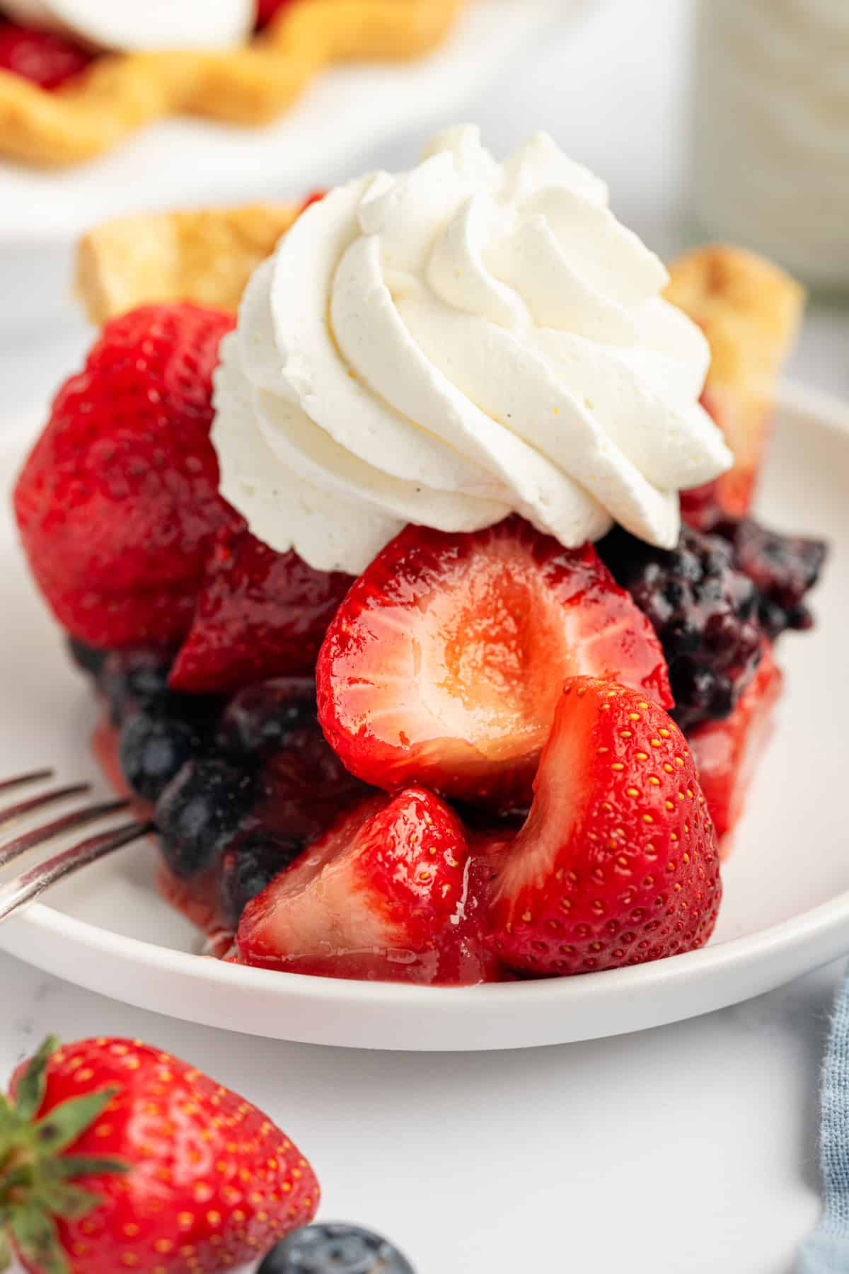 Berry Pie A slice of berry pie topped with whipped cream sits on a white plate, featuring whole strawberries and blueberries.