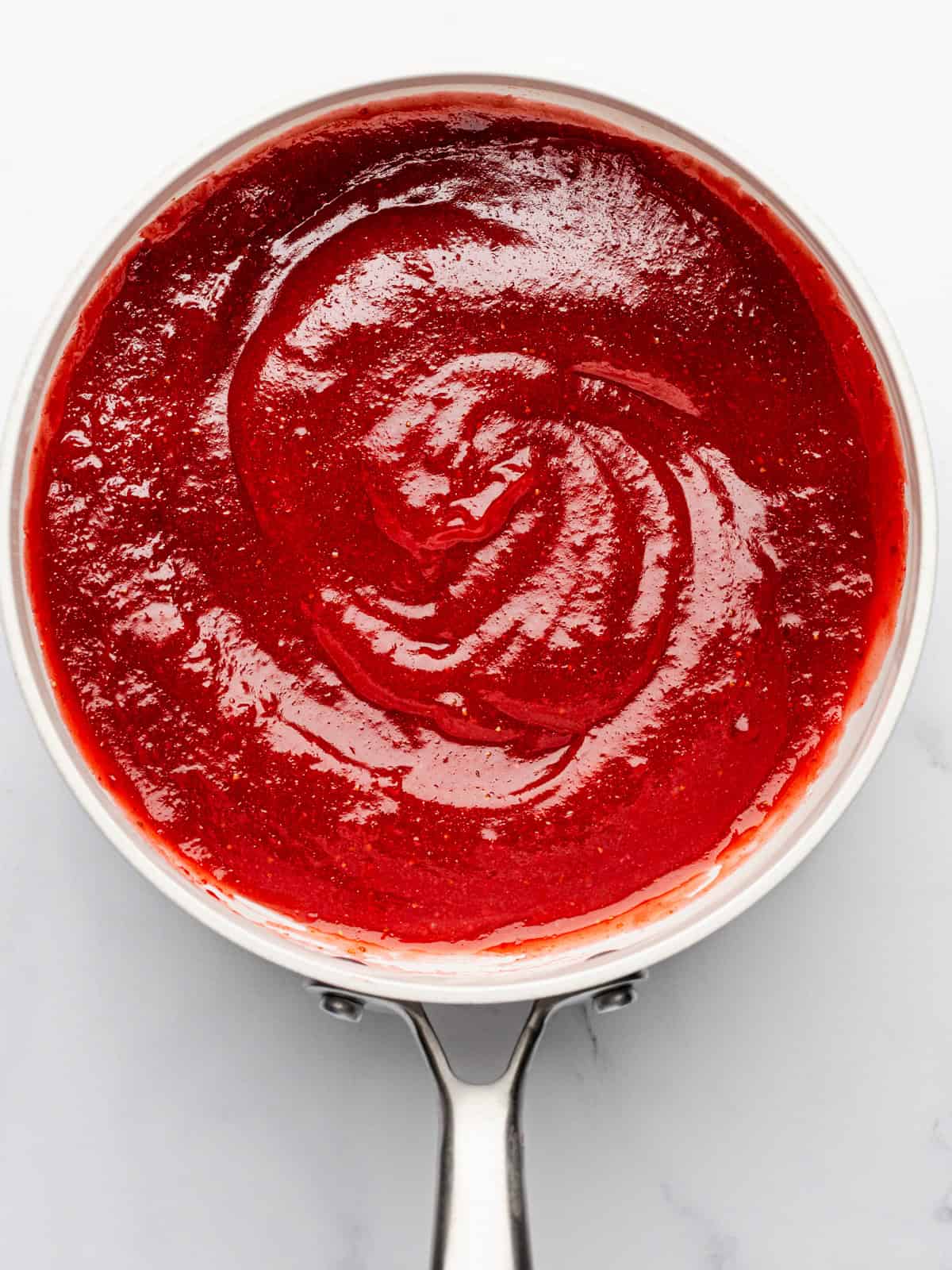 Berry Pie A silver saucepan filled with smooth, glossy red sauce, swirled in the center, sits on a white surface.