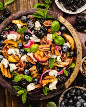 A vibrant salad in a wooden bowl with blackberries, blueberries, peaches, pecans, red onions, fresh mint, basil leaves, and crumbled cheese, surrounded by small bowls of berries and pecans on a rustic wooden table.