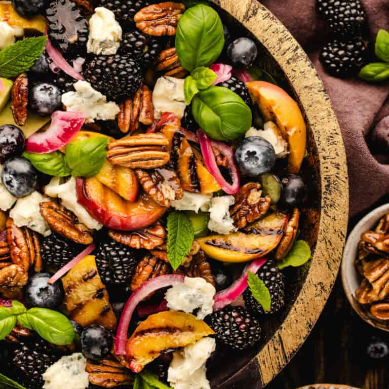 A colorful salad in a rustic wooden bowl, featuring blackberries, blueberries, grilled peach slices, pecans, red onion, fresh basil, mint leaves, and crumbled cheese.