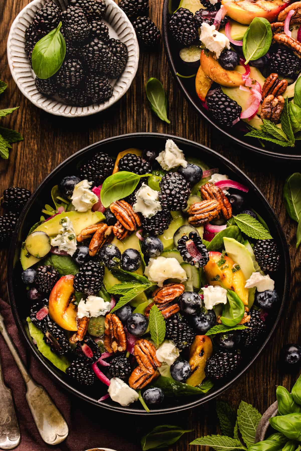 Blackberry Peach Salad with Avocado, Gorgonzola, Blueberries, Spinach, Cucumber, Pickled Onions, and Maple Pecans. A vibrant salad with blackberries, blueberries, peach slices, pecans, and chunks of cheese on greens. Fresh mint and basil leaves garnish the dish. A small bowl of blackberries sits nearby on a wooden table.