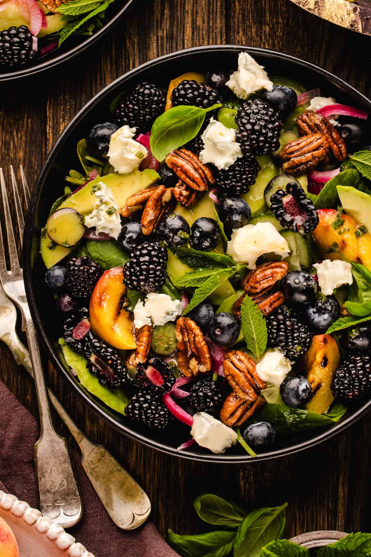 Blackberry Peach Salad with Avocado, Gorgonzola, Blueberries, Spinach, Cucumber, Pickled Onions, and Maple Pecans. A fresh salad with blackberries, blueberries, pecans, peach slices, greens, red onions, and crumbled cheese, served in a black bowl on a wooden table with a fork and knife nearby.