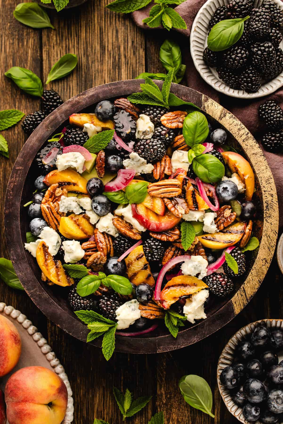 Blackberry Peach Salad with Avocado, Gorgonzola, Blueberries, Spinach, Cucumber, Pickled Onions, and Maple Pecans. A wooden bowl filled with salad made of blackberries, blueberries, peach slices, pecans, red onion, crumbled cheese, and fresh mint leaves, placed on a wooden table with extra fruit and herbs scattered around.