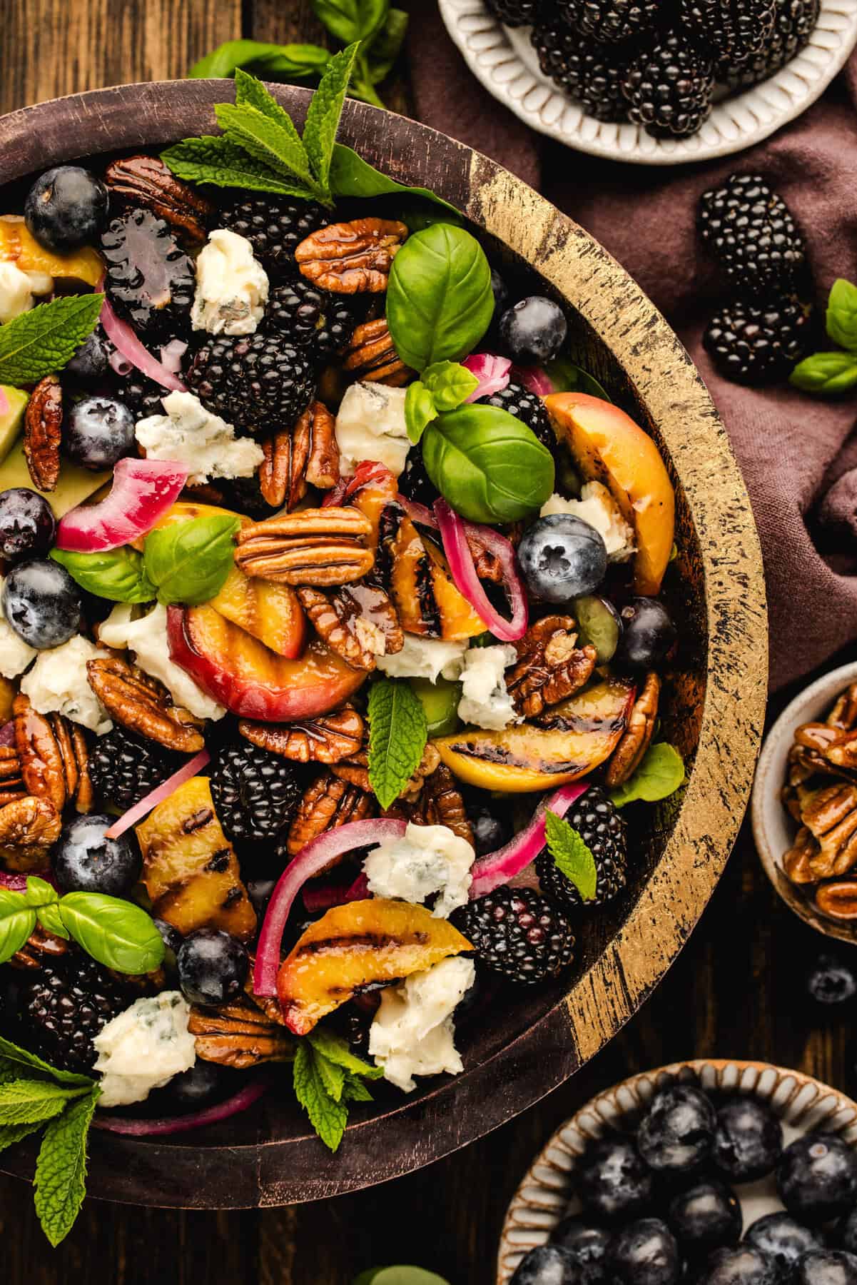 Blackberry Peach Salad with Avocado, Gorgonzola, Blueberries, Spinach, Cucumber, Pickled Onions, and Maple Pecans. A wooden bowl filled with a colorful salad of blackberries, blueberries, peaches, pecans, gorgonzola cheese, pickled onions, and fresh basil and mint leaves. Small bowls of blackberries and pecans sit nearby.
