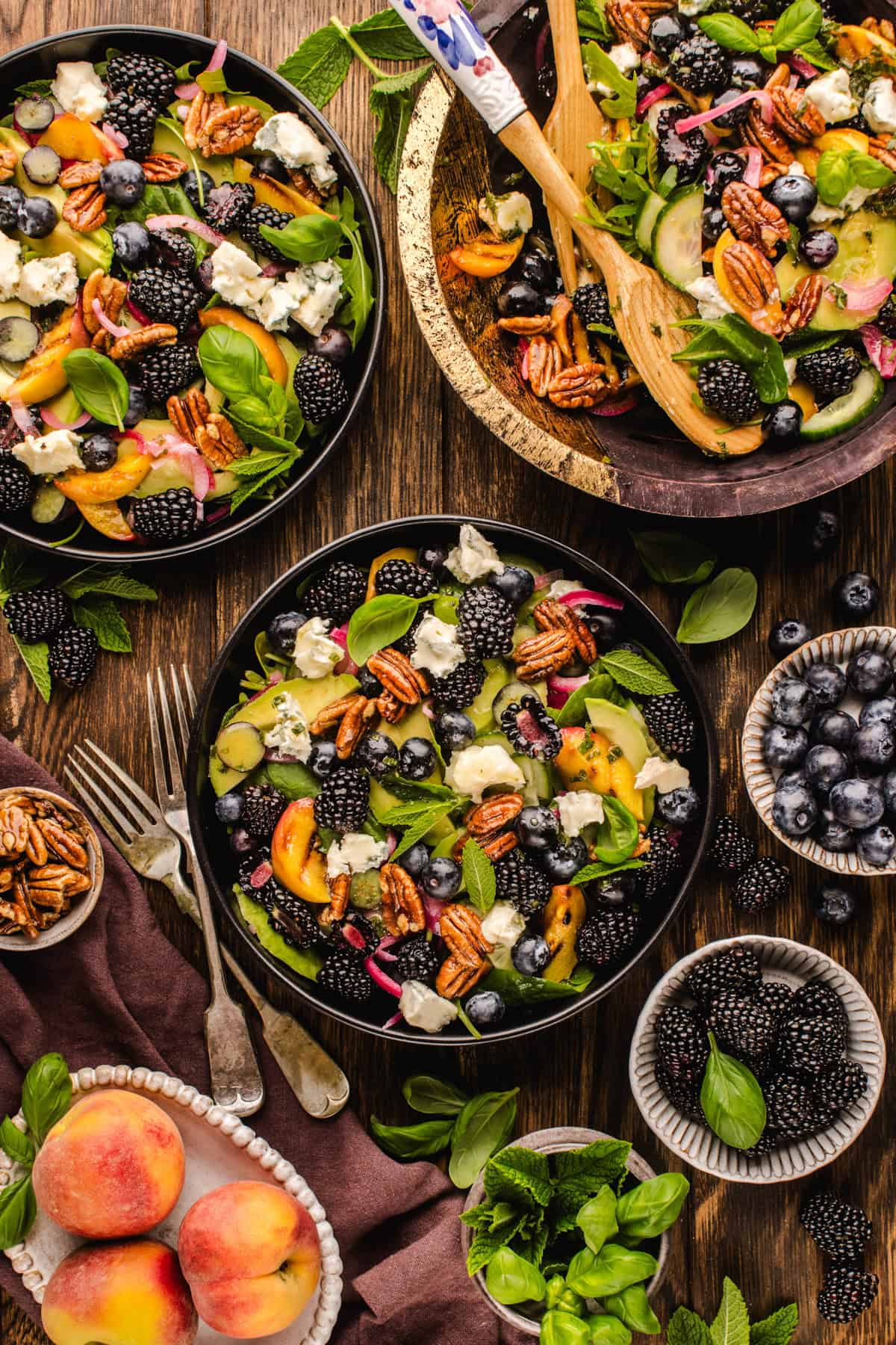 Blackberry Peach Salad with Avocado, Gorgonzola, Blueberries, Spinach, Cucumber, Pickled Onions, and Maple Pecans. Overhead view of plates and bowls filled with a colorful salad of blackberries, blueberries, peaches, pecans, cheese crumbles, red onions, cucumber, and fresh basil on a wooden table, surrounded by fresh ingredients.