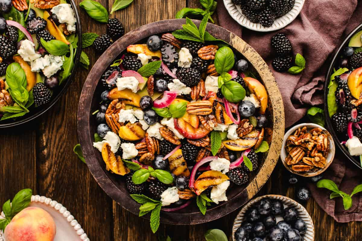 Blackberry Peach Salad with Avocado, Gorgonzola, Blueberries, Spinach, Cucumber, Pickled Onions, and Maple Pecans. A colorful salad in a wooden bowl with blackberries, blueberries, peach slices, pecans, gorgonzola cheese, red onions, and fresh basil leaves, surrounded by small bowls of pecans and blueberries on a wooden table.
