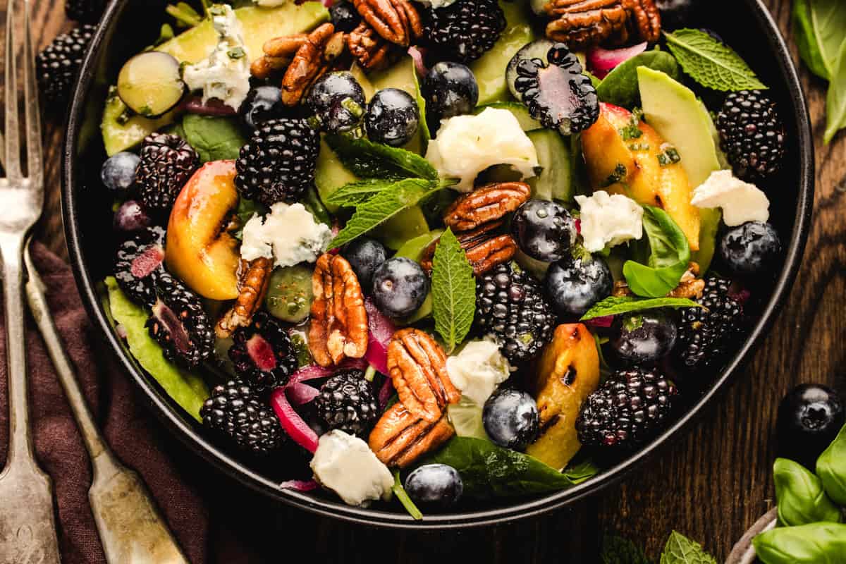 Blackberry Peach Salad with Avocado, Gorgonzola, Blueberries, Spinach, Cucumber, Pickled Onions, and Maple Pecans. A close-up of a vibrant salad with spinach, blackberries, blueberries, peach slices, pecans, red onion, fresh mint, and crumbled cheese in a black bowl on a wooden table.