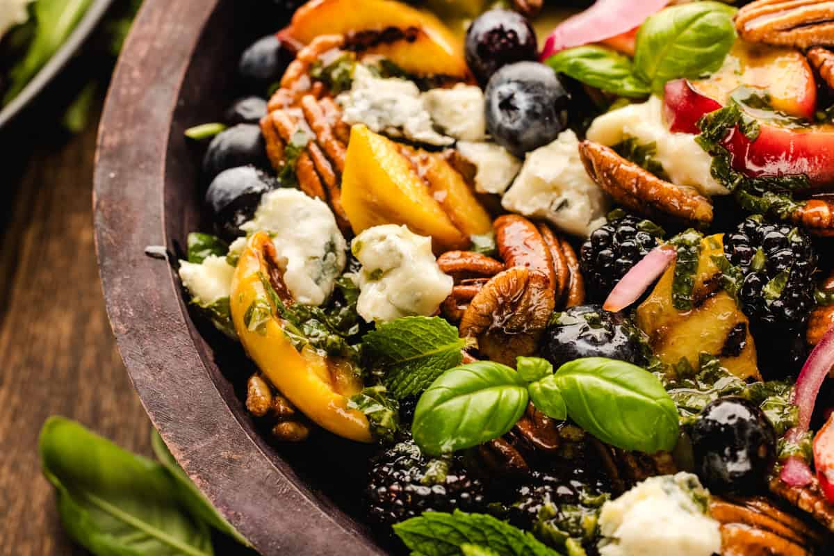 Blackberry Peach Salad with Avocado, Gorgonzola, Blueberries, Spinach, Cucumber, Pickled Onions, and Maple Pecans. A close-up of a colorful salad with fresh peaches, blueberries, blackberries, pecans, gorgonzola cheese crumbles, red onion, and basil leaves in a wooden bowl.