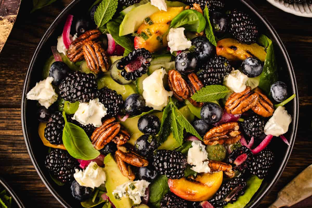 Blackberry Peach Salad with Avocado, Gorgonzola, Blueberries, Spinach, Cucumber, Pickled Onions, and Maple Pecans. A colorful salad with pecans, blackberries, blueberries, peach slices, fresh mint, basil leaves, red onion, and crumbled white cheese on a bed of mixed greens.