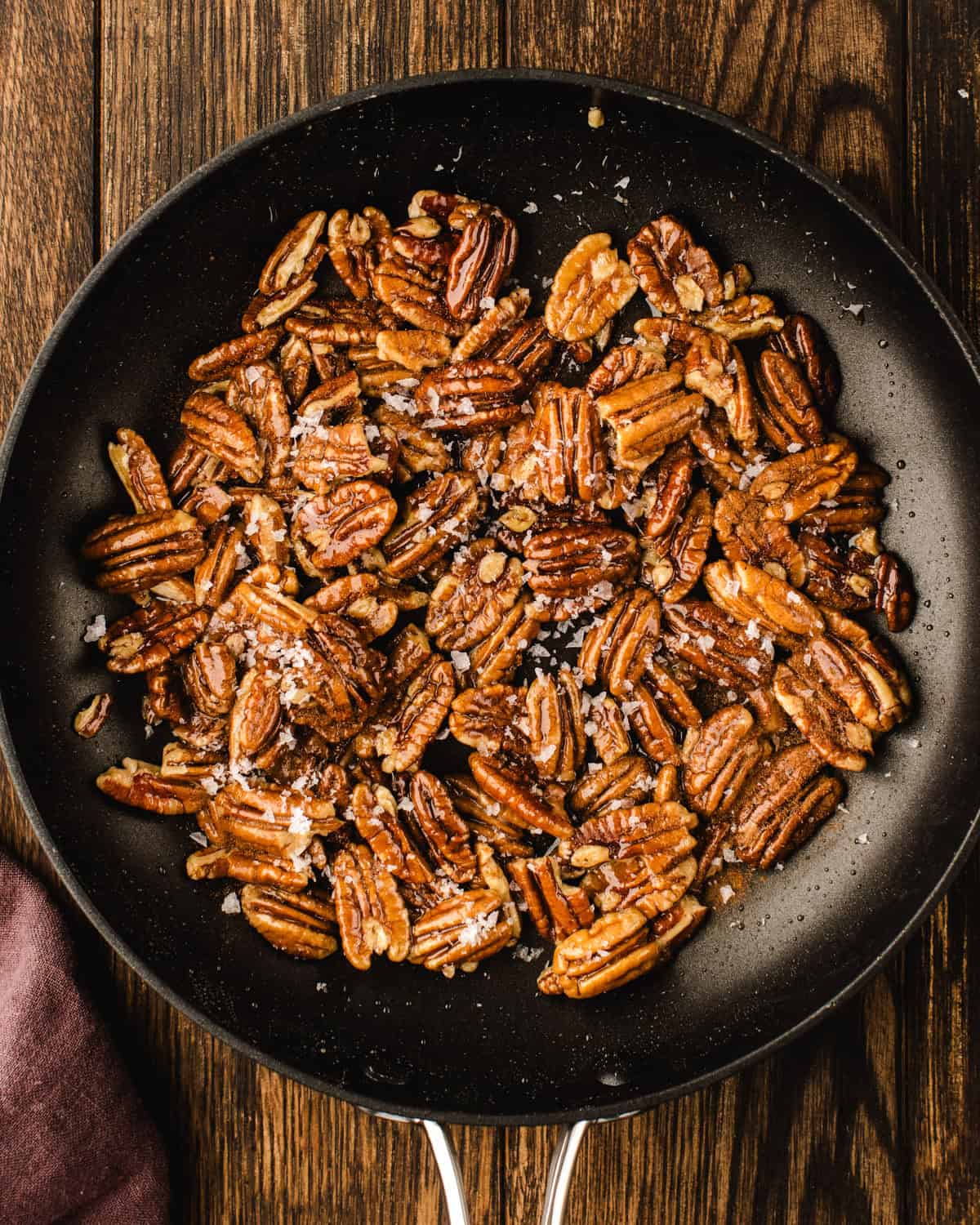 Candied Pecans for Blackberry Peach Salad. A black skillet (filled with toasted pecans, sprinkled with coarse salt and cinnamon, mixed with maple syrup) sits on a wooden surface.