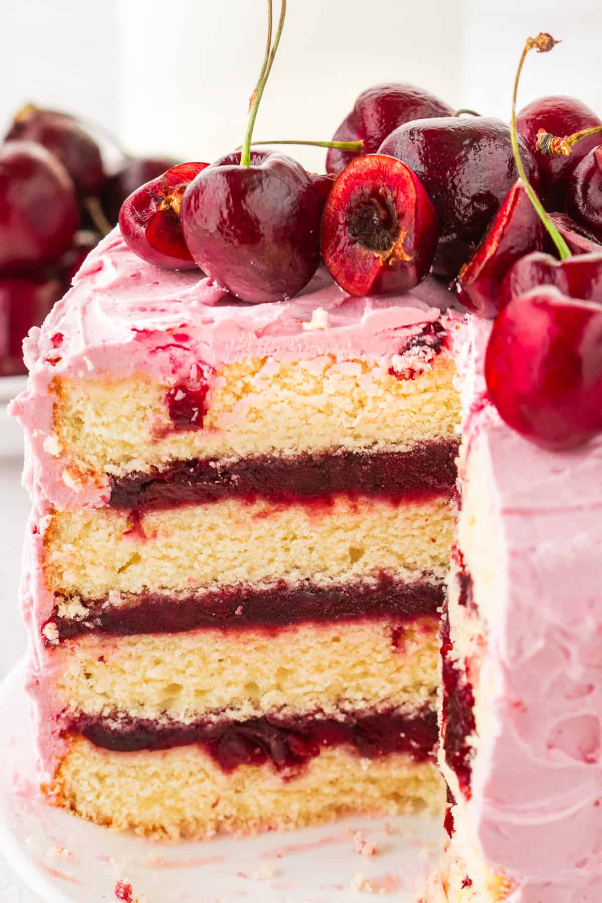 Cherry Layer Cake Three-layer vanilla cake with cherry filling and pink frosting, topped with whole and halved cherries; a large slice is cut out, revealing the cake's interior.
