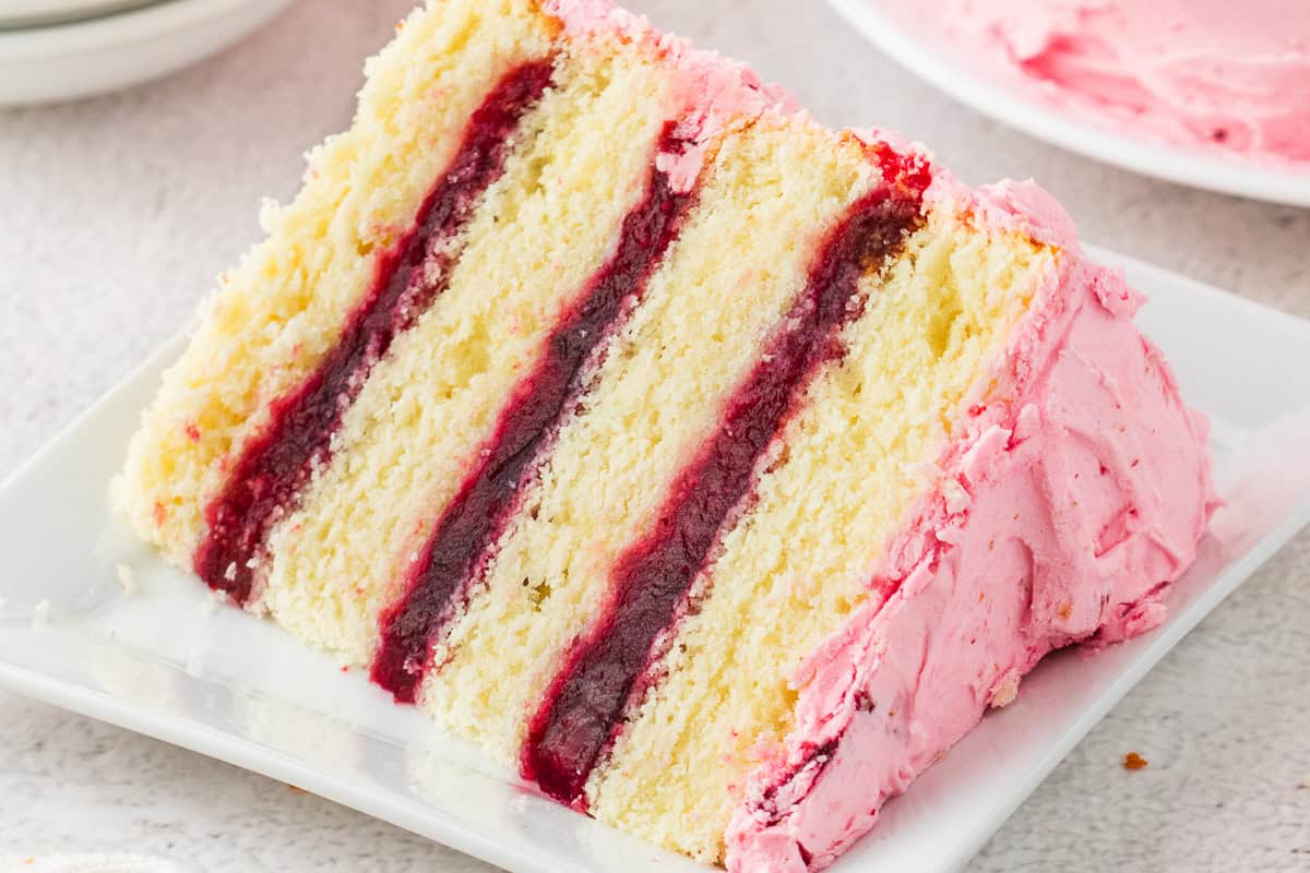 Cherry Layer Cake A slice of layered cake with pink frosting and dark red jam filling, served on a white plate.
