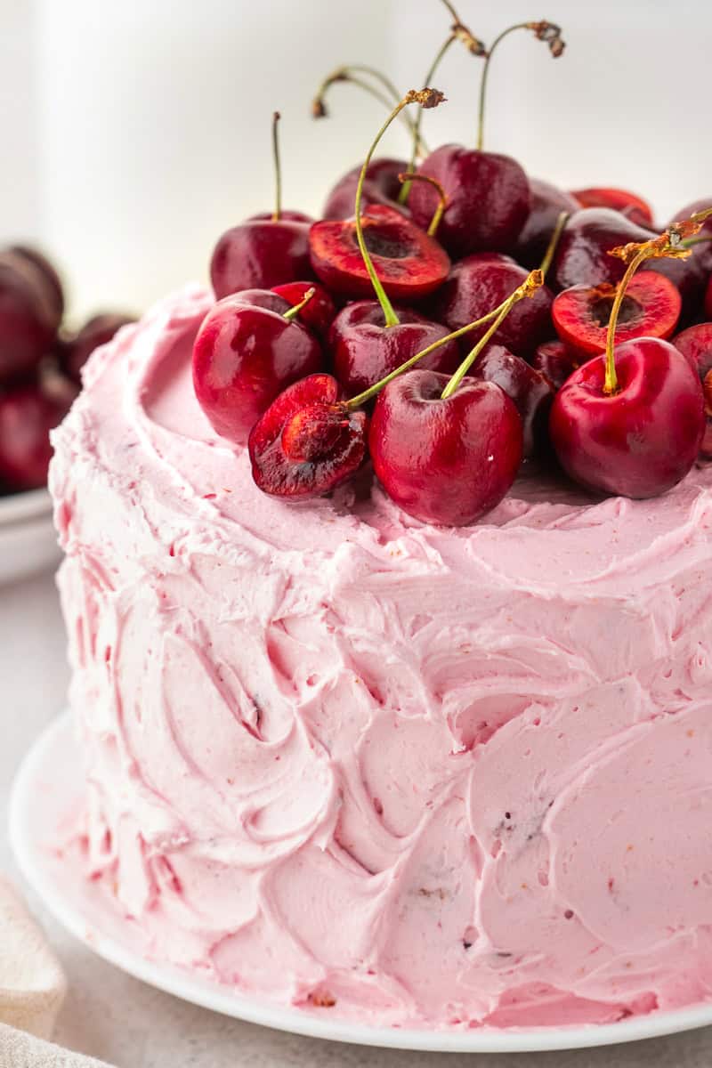 Cherry Layer Cake A round cake with swirled pink frosting, topped with a generous pile of fresh, whole and halved cherries on stems, sits on a white plate.