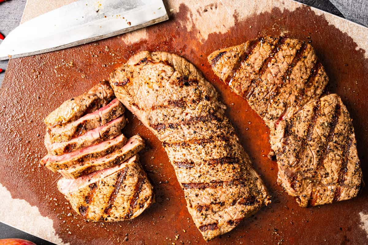 Thai Beef Salad (Step-by-Step Photos) Three grilled steaks rest on a wooden cutting board, one sliced, showing a pink center.