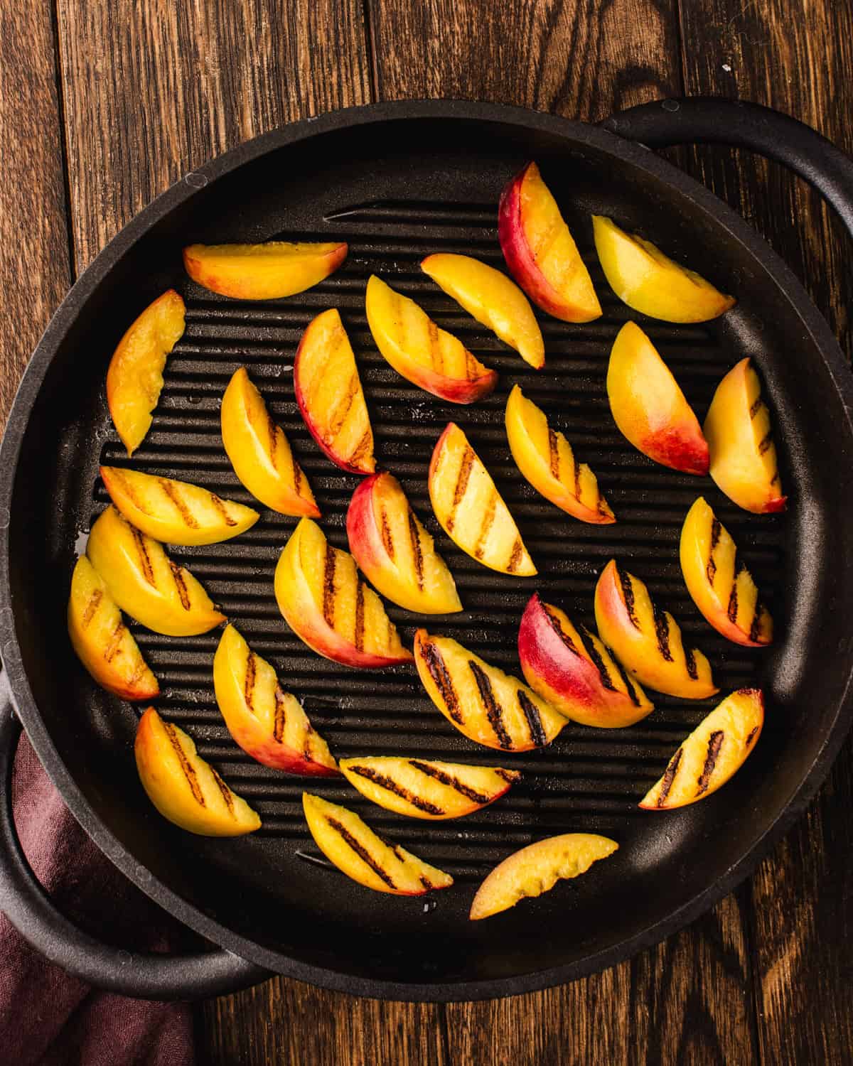 Grilled Peaches for Blackberry Peach Salad. Slices of fresh peaches with visible grill marks arranged on a black grill pan, set on a rustic wooden surface.