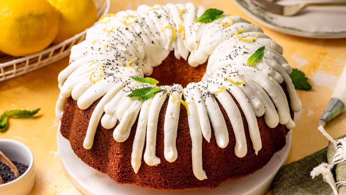 Poppy Seed Lemon Bundt Cake Lemon poppy seed bundt cake with white icing drizzled on top, garnished with lemon zest, poppy seeds, and mint leaves, sits on a round white plate.