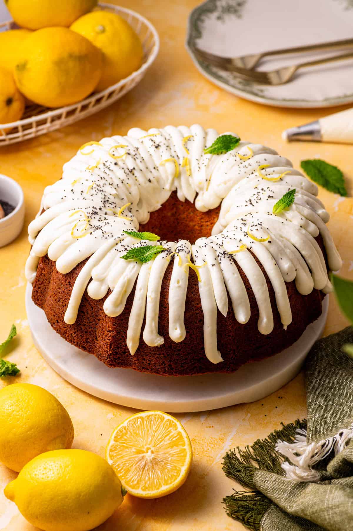 Poppy Seed Lemon Bundt Cake A lemon bundt cake with white icing, garnished with lemon zest, poppy seeds, and mint leaves, sits on a white plate.