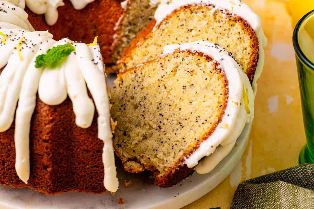 Poppy Seed Lemon Bundt Cake A close-up of a sliced lemon poppy seed bundt cake with white icing drizzled on top, garnished with poppy seeds and a mint leaf.