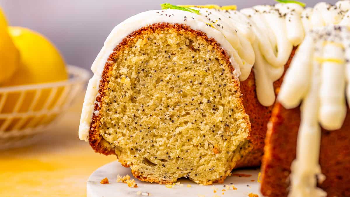 Poppy Seed Lemon Bundt Cake A close-up of a sliced lemon poppy seed bundt cake with white icing drizzled on top, sitting on a marble surface.