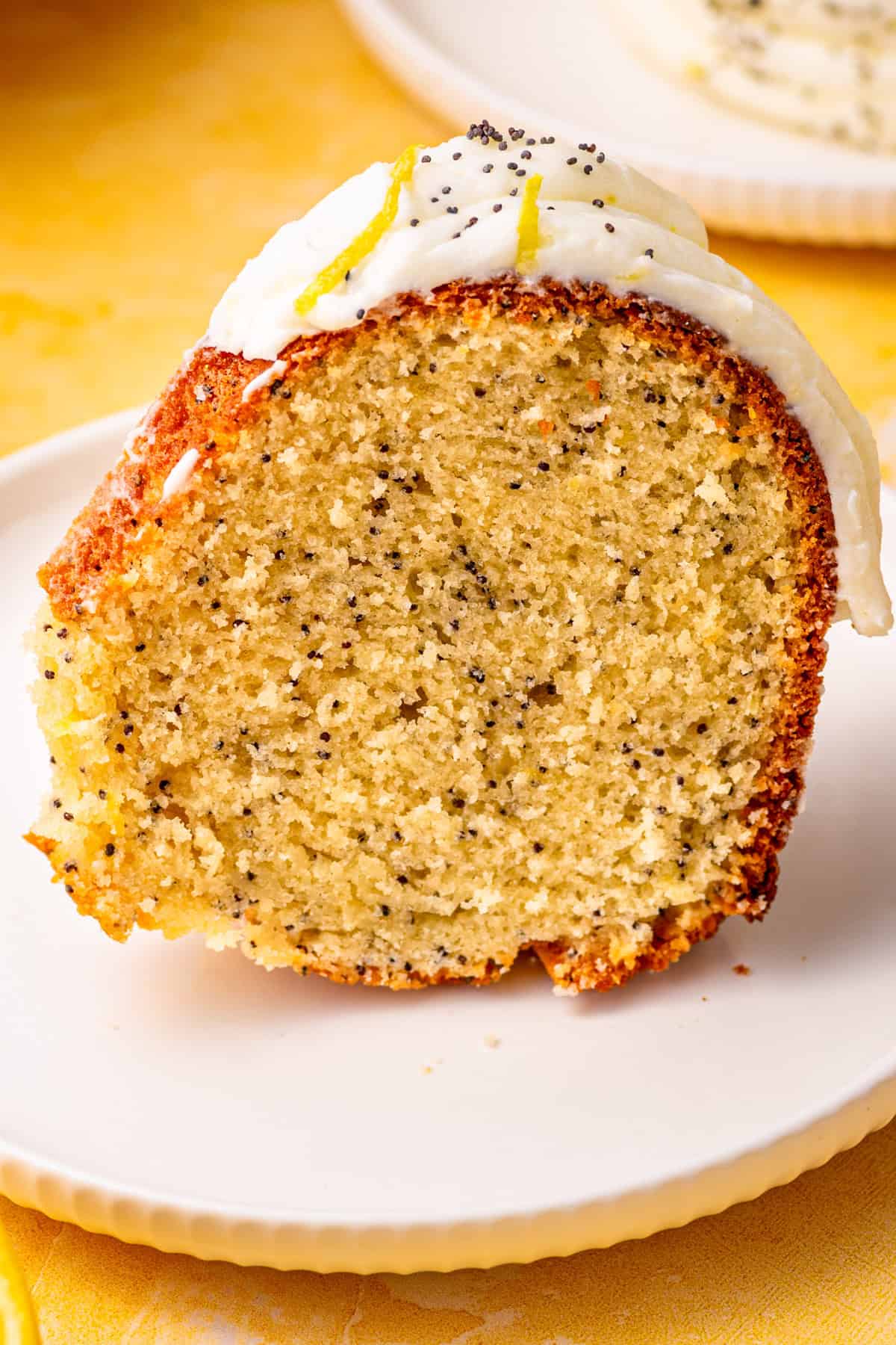 Poppy Seed Lemon Bundt Cake A close-up of a slice of lemon poppy seed bundt cake with white icing, lemon zest, and poppy seeds on top, served on a white plate.