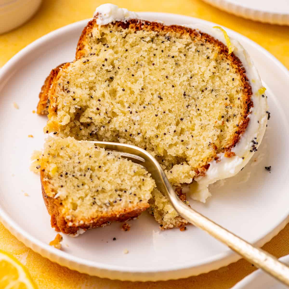 Poppy Seed Lemon Bundt Cake A close-up of a slice of lemon poppy seed cake with white icing on a plate. A fork is cutting a piece from the cake.