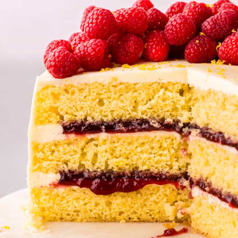 A close-up of a layered lemon raspberry cake with white frosting, raspberry jam filling, and fresh raspberries piled on top. A slice has been cut out, revealing the cakes moist, fluffy texture and vibrant layers.