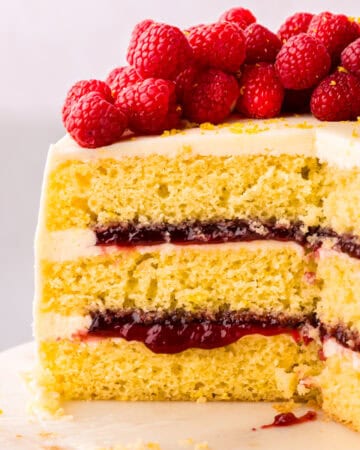 A layered vanilla sponge lemon raspberry cake with white frosting, filled with red jam, topped with fresh raspberries, and a slice is cut out, showing the inside.