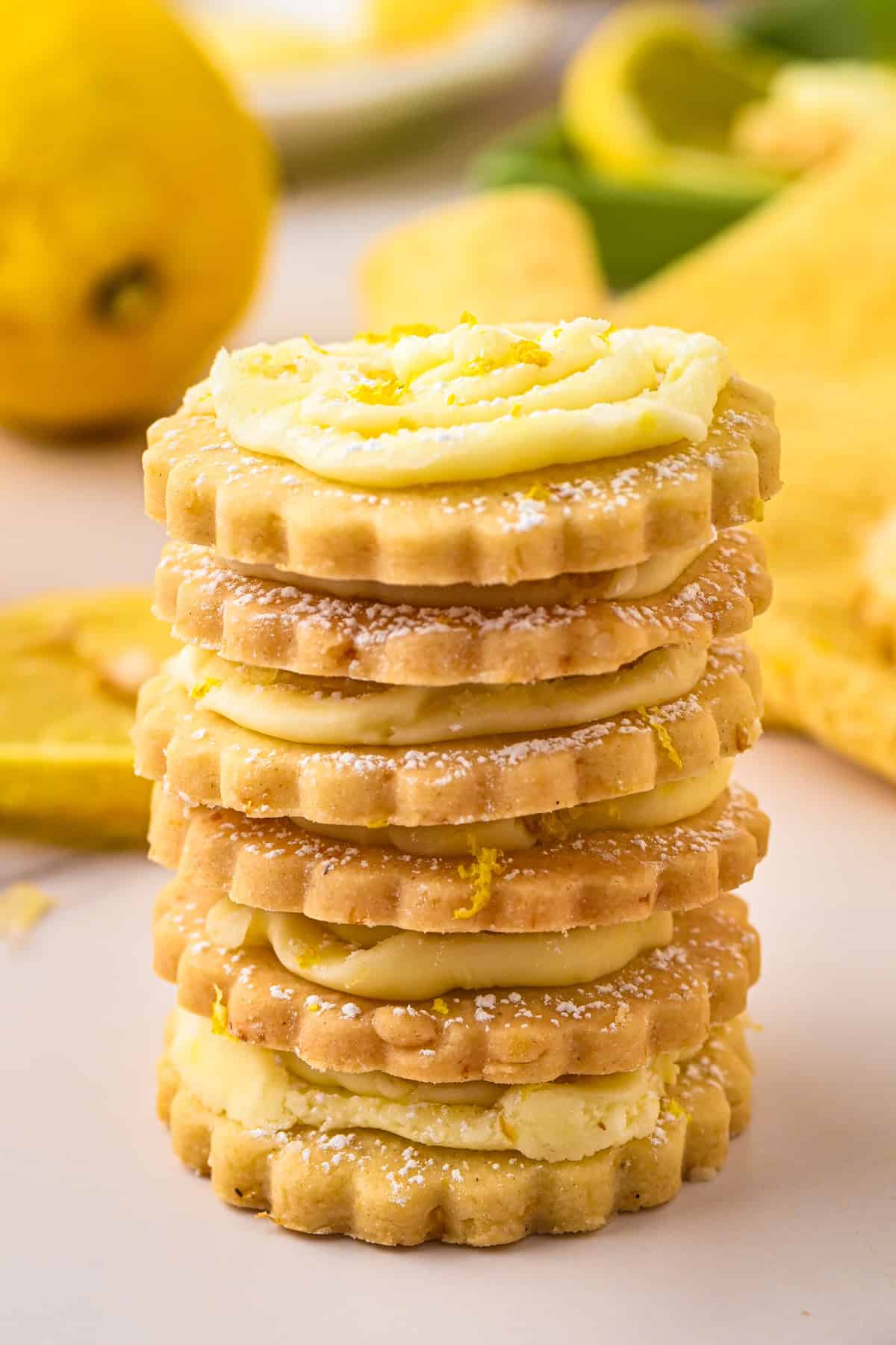 Lemon Shortbread Cookies A stack of lemon cream sandwich cookies with scalloped edges, filled with frosting and topped with powdered sugar, sits on a light surface with lemons in the background.