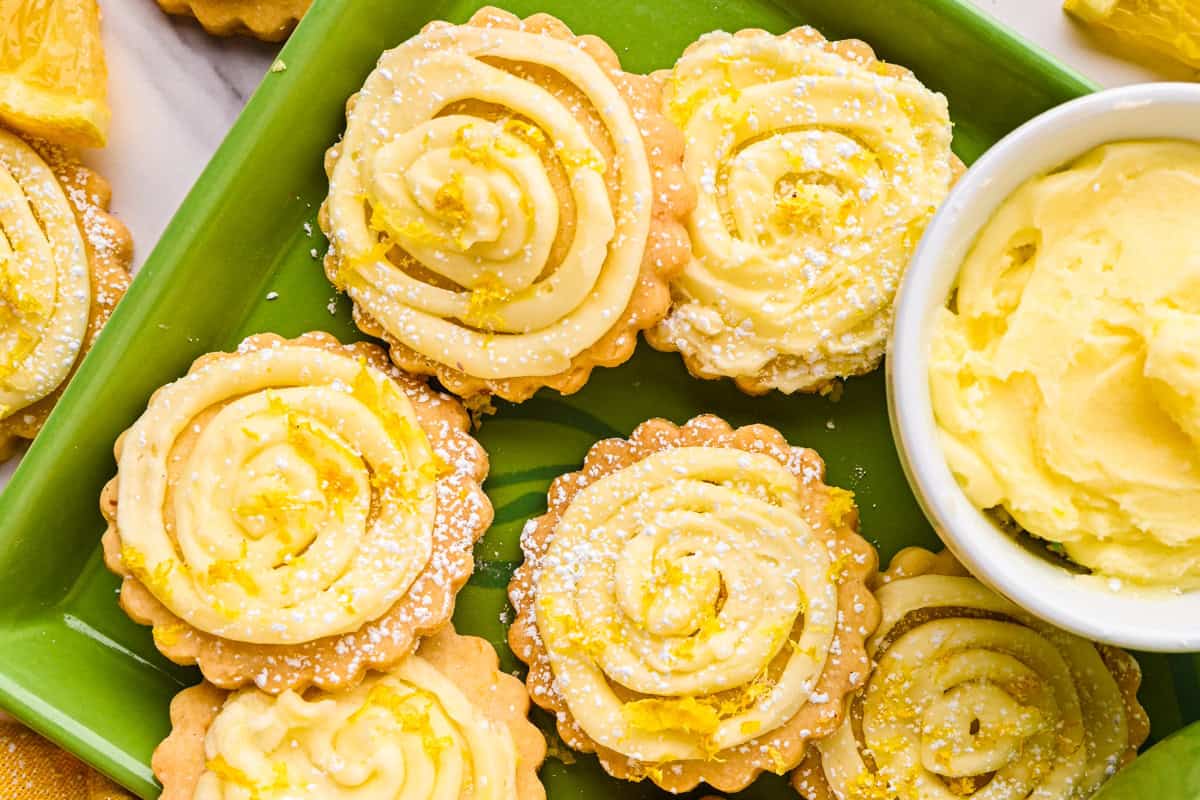 Lemon Shortbread Cookies A green plate with lemon cookies topped with swirled icing and lemon zest, dusted with powdered sugar.