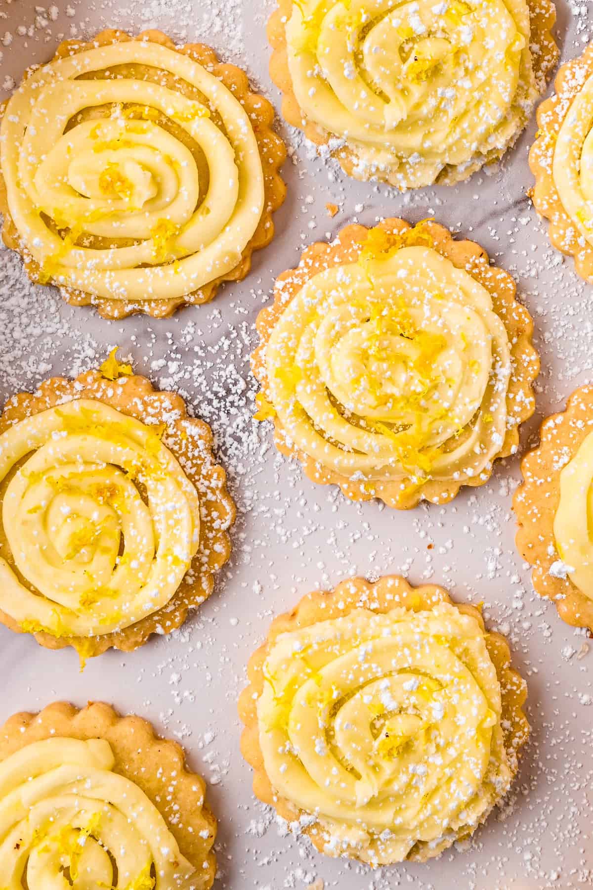 Lemon Shortbread Cookies Round cookies topped with swirls of yellow frosting and sprinkled with powdered sugar and lemon zest, arranged on a light surface.