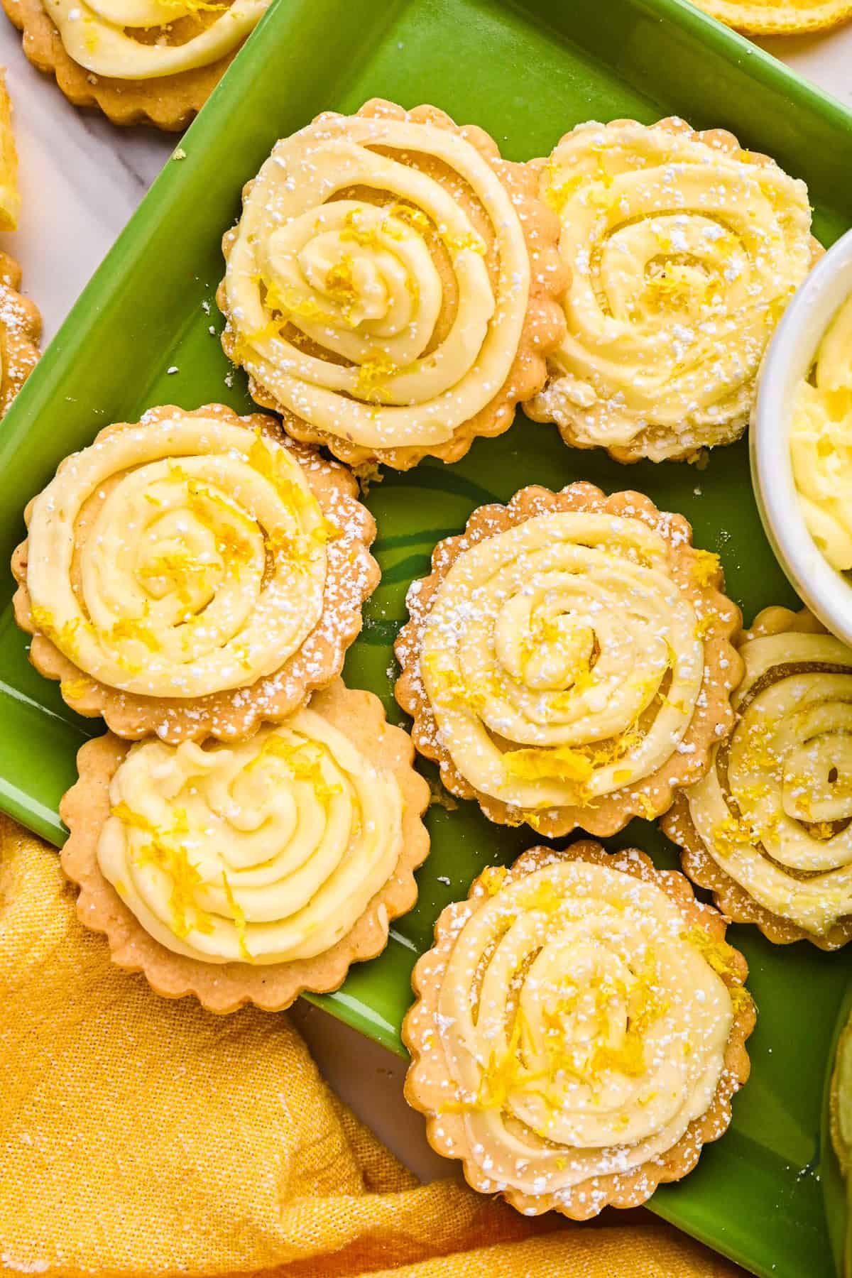 Lemon Shortbread Cookies Round lemon shortbread cookies with swirled yellow lemon buttercream and dusted with powdered sugar are arranged on a green tray.