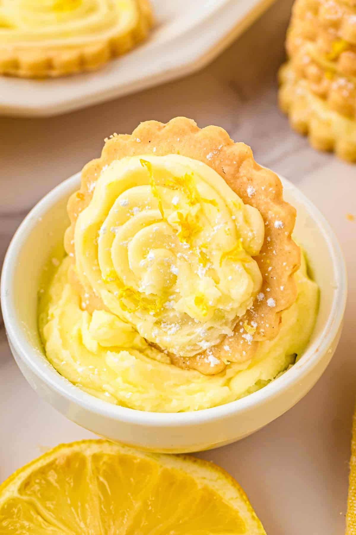 Lemon Shortbread Cookies A cookie with piped lemon frosting and lemon zest rests upright in a small bowl of creamy lemon frosting, with a lemon slice nearby.