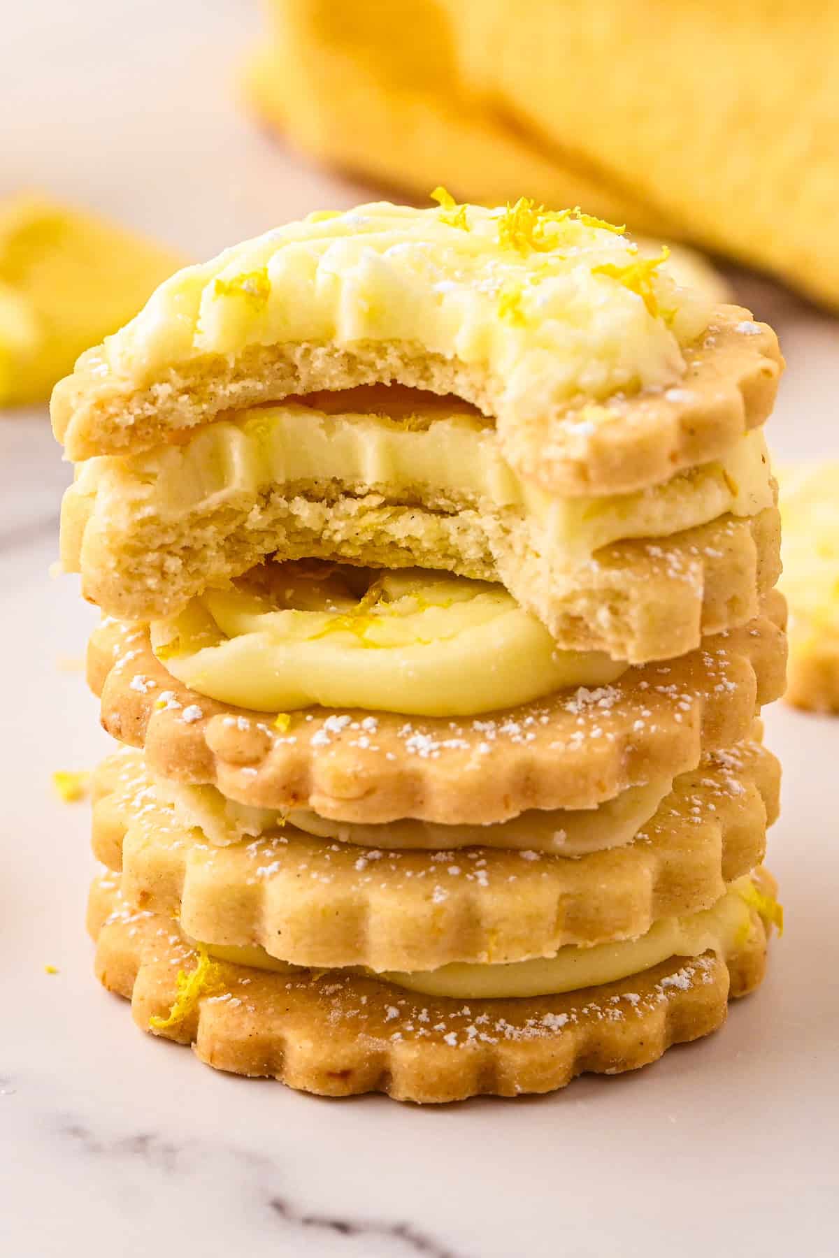 Lemon Shortbread Cookies A stack of five lemon cream sandwich cookies, with the top cookie partially bitten to reveal the creamy filling. Cookie edges are scalloped, and lemon zest is sprinkled on top and around them.