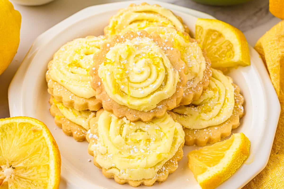 Lemon Shortbread Cookies A white plate filled with lemon-flavored cookies topped with yellow frosting and powdered sugar, surrounded by lemon wedges.