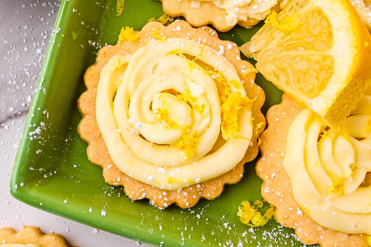 Lemon Shortbread Cookies Close-up of lemon shortbread cookies with swirled lemon frosting, garnished with lemon zest and a lemon wedge, on a green plate. Powdered sugar is sprinkled on top and around the tarts.
