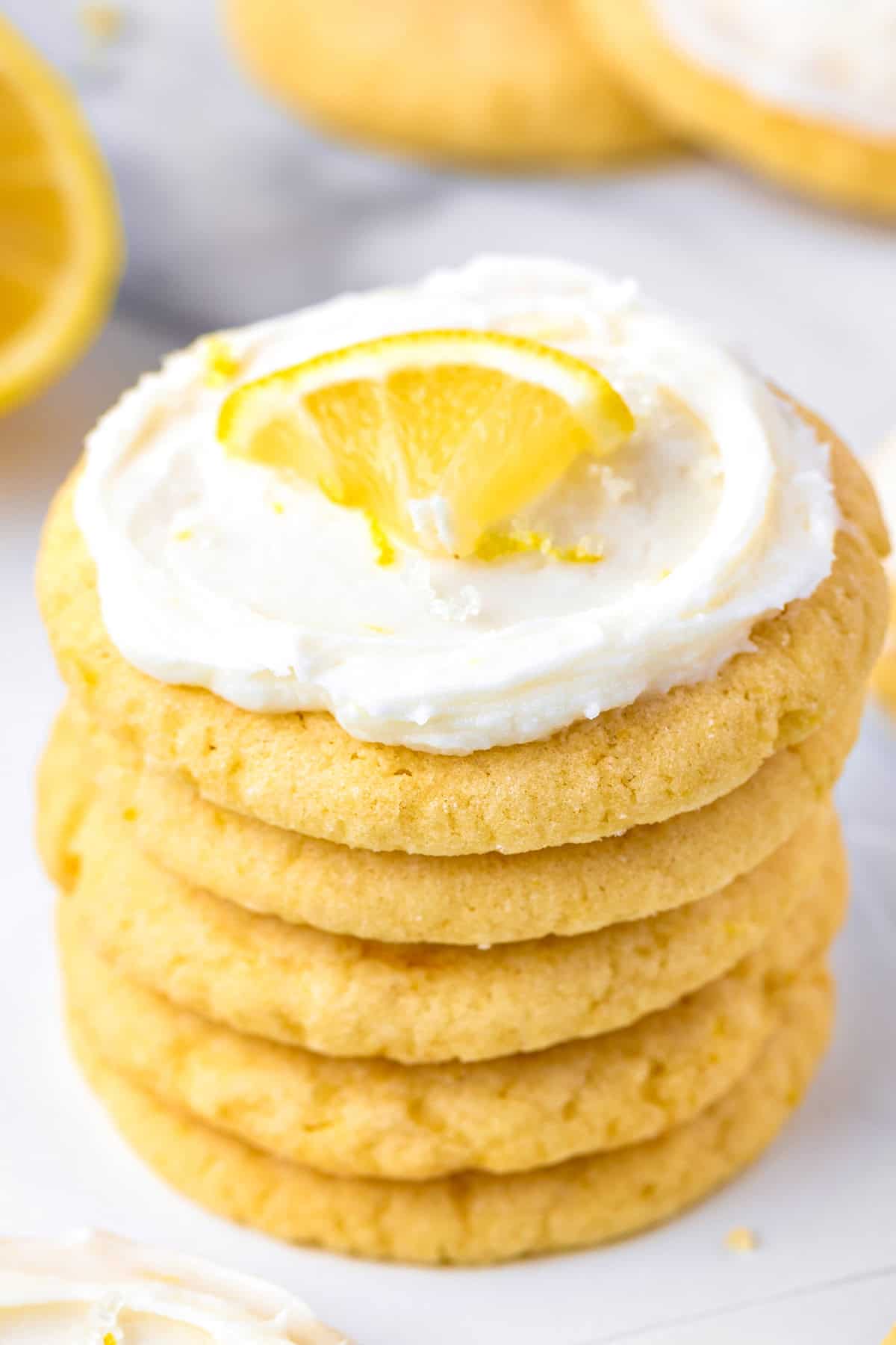 Lemon Sugar Cookies A stack of lemon cookies with white frosting, topped with a small lemon wedge, sits on a white surface.