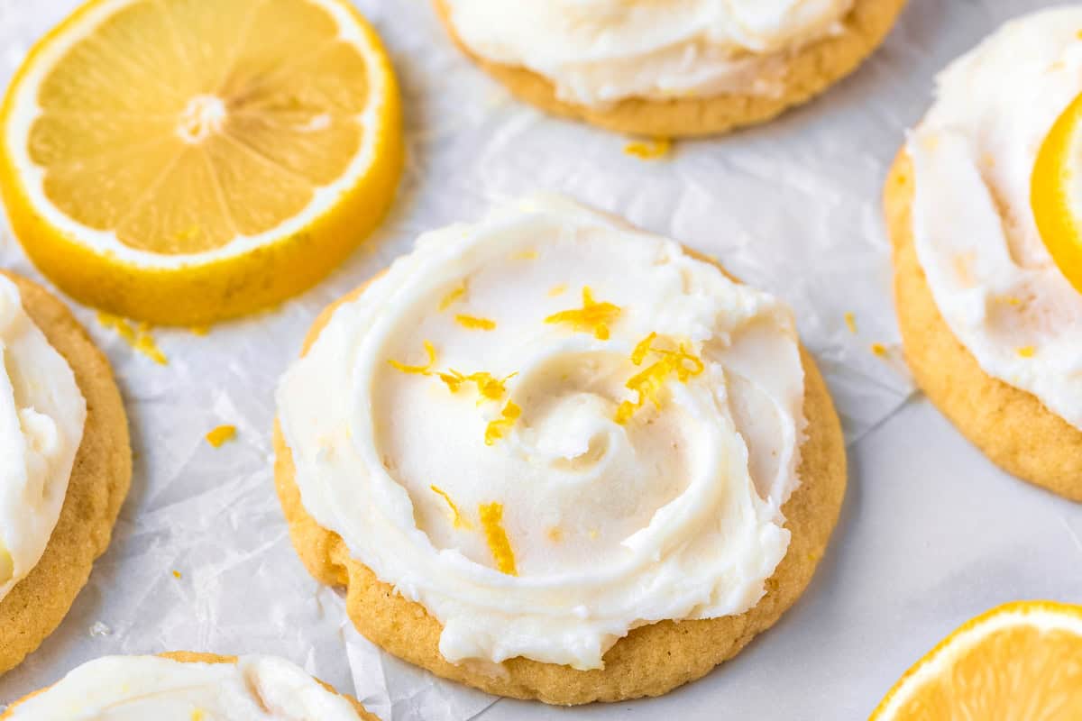 Lemon Sugar Cookies A close-up of a lemon sugar cookie with creamy white frosting and lemon zest on top, surrounded by lemon slices on parchment paper.