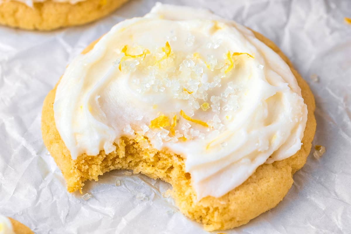 Lemon Sugar Cookies A frosted sugar cookie with a bite taken out, topped with white icing, coarse sugar, and lemon zest, sits on crinkled parchment paper.