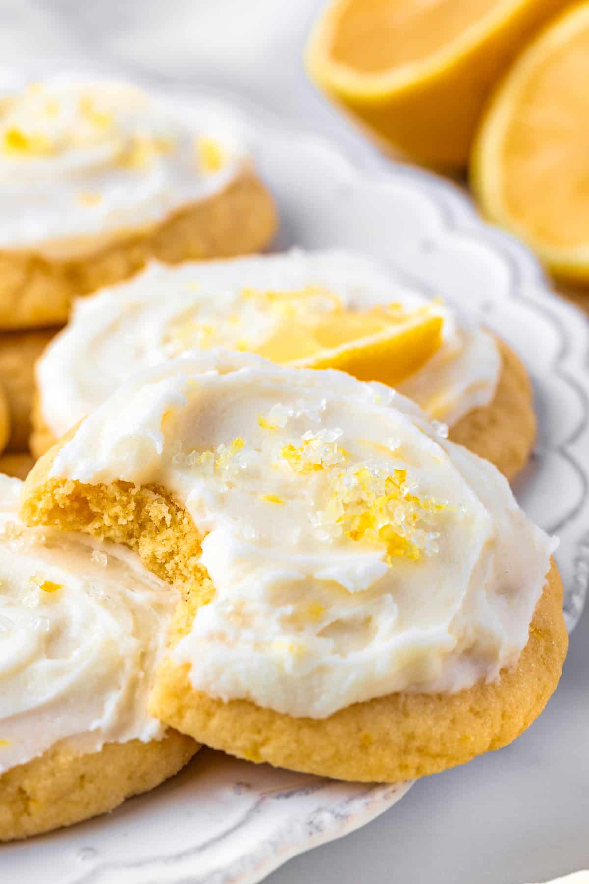 Lemon Sugar Cookies Frosted lemon cookies on a plate, topped with lemon zest and sugar crystals. One cookie has a bite taken out, and lemon halves are visible in the background.