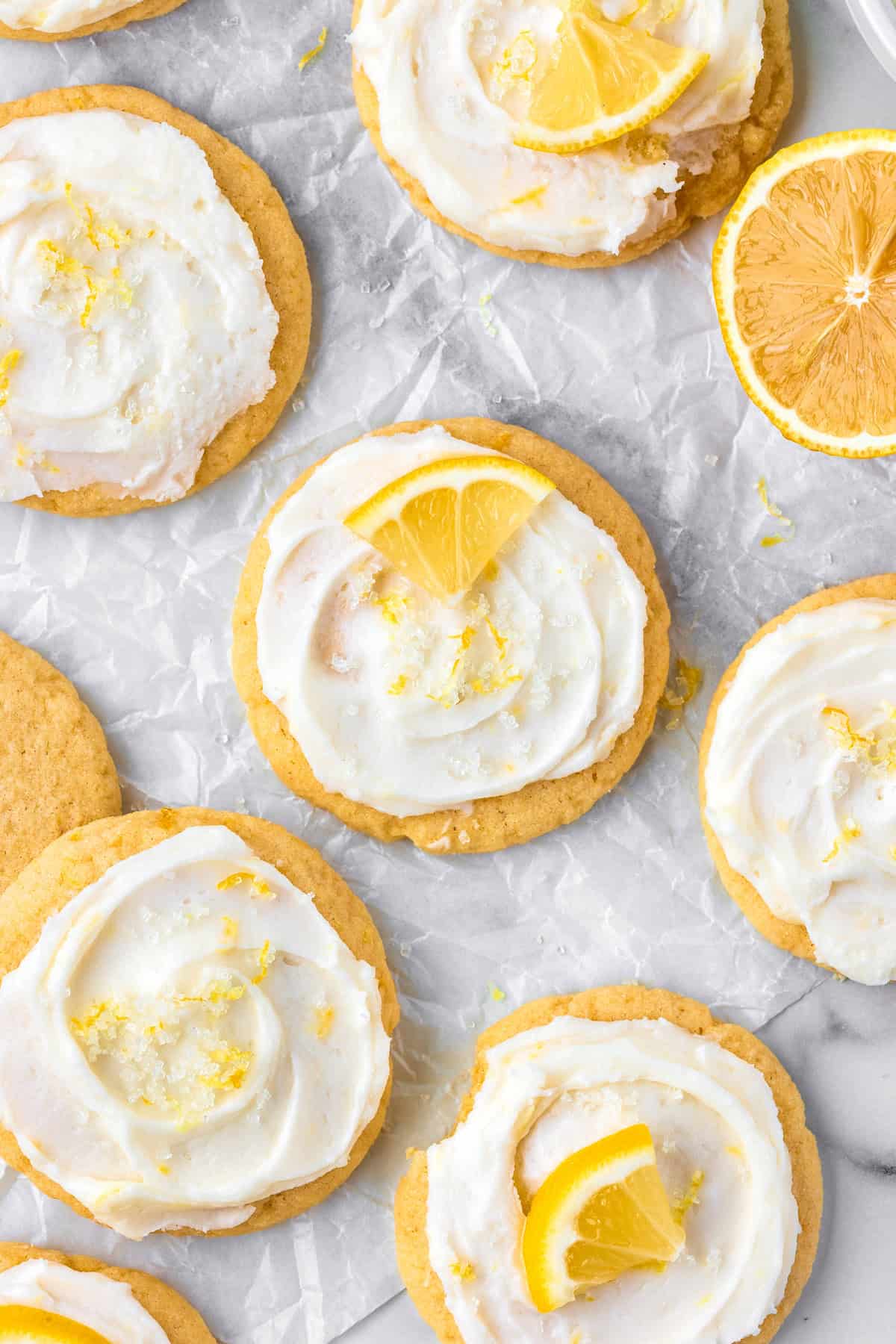 Lemon Sugar Cookies Lemon cookies with white icing and lemon slices on top are arranged on parchment paper. Slices of lemon and cookie crumbs are visible around the cookies.
